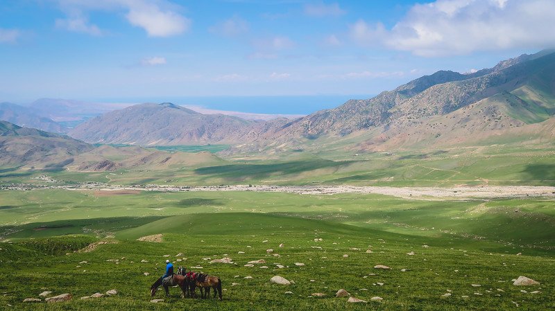 A horse rider pausing on a grassy hillside with sweeping views of green valleys, rugged mountains, and a distant lake during the 3-Day Panorama Trek in Kyrgyzstan