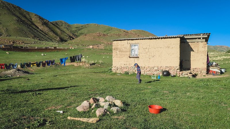 A small mud-brick homestay in a remote Kyrgyz mountain valley, with laundry hanging on a clothesline, a child playing outside, and green hills in the background during the second night of the 3-Day Panorama Trek
