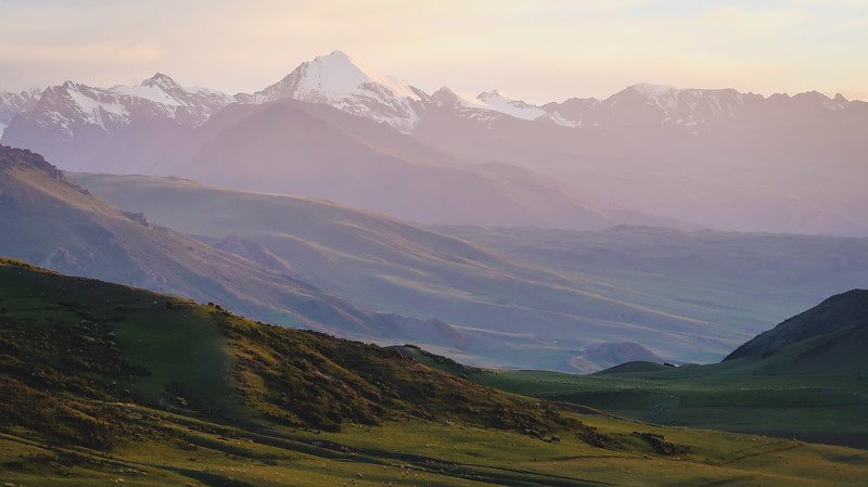 Snow-capped peaks of the Tian Shan Mountains glowing in soft pastel light during a stunning sunset over rolling green valleys on the 3-Day Panorama Trek in Kyrgyzstan