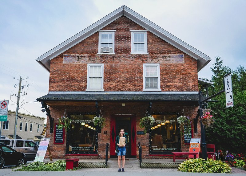 That Backpacker Audrey Bergner standing outside La Rumeur Affamée, a gourmet food shop housed in a charming red-brick heritage building in Sutton, Quebec, holding a brown paper bag of local treats
