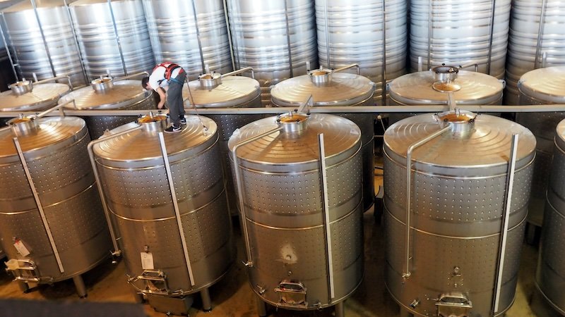 Behind-the-scenes tour at La Vinyeta winery in Costa Brava, Spain, showing a worker with a safety harness inspecting large stainless steel wine storage tanks, highlighting the meticulous process and equipment used in modern winemaking