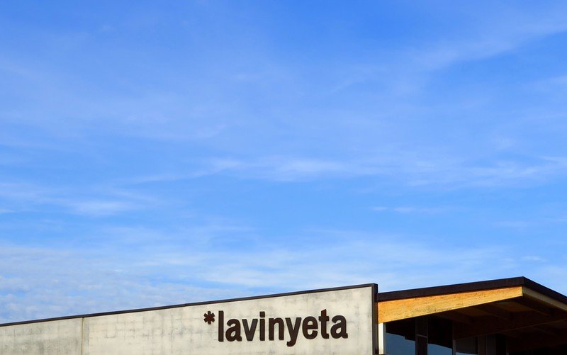 Modern exterior of La Vinyeta winery in Costa Brava, Spain, featuring its bold signage against a bright blue sky, marking the start of a vineyard tour that includes grape stomping, wine tasting, and discovering Catalonia’s wine culture