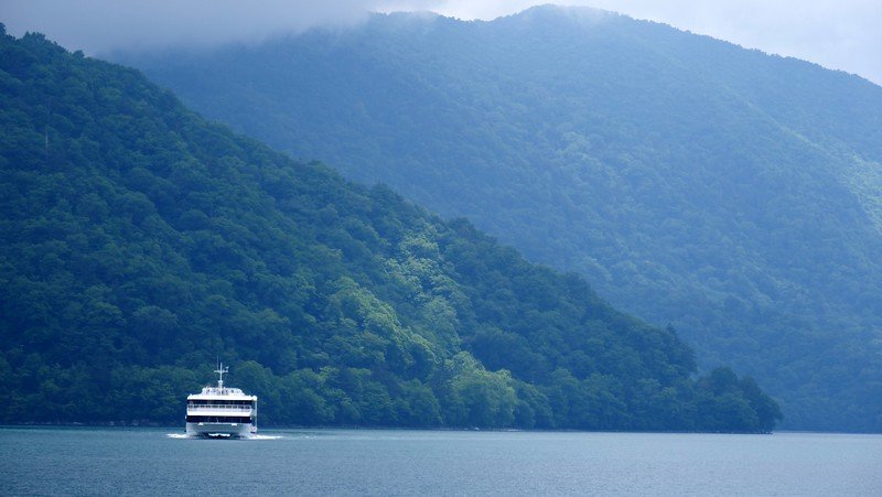 Scenic boat cruising across the tranquil waters of Lake Chuzenji surrounded by lush green forested mountains and low-hanging mist in Nikko, Japan