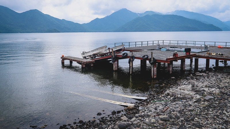 Wooden docks extending into the calm waters of Lake Chuzenji surrounded by misty blue mountain ranges in Nikko, Japan, with small boats and fishing gear resting on the pier
