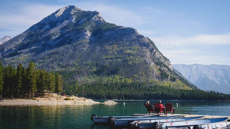 Tranquil turquoise waters of Lake Minnewanka reflecting a towering forested mountain, with red Adirondack chairs on a dock and small boats moored nearby during our Canadian Rockies road trip