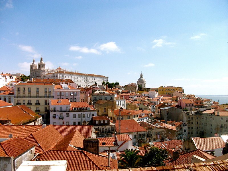 Classic Lisbon view over the Alfama district with red rooftops, narrow winding streets, and landmarks like the São Vicente de Fora Monastery and National Pantheon under a sunny blue sky overlooking the Tagus River
