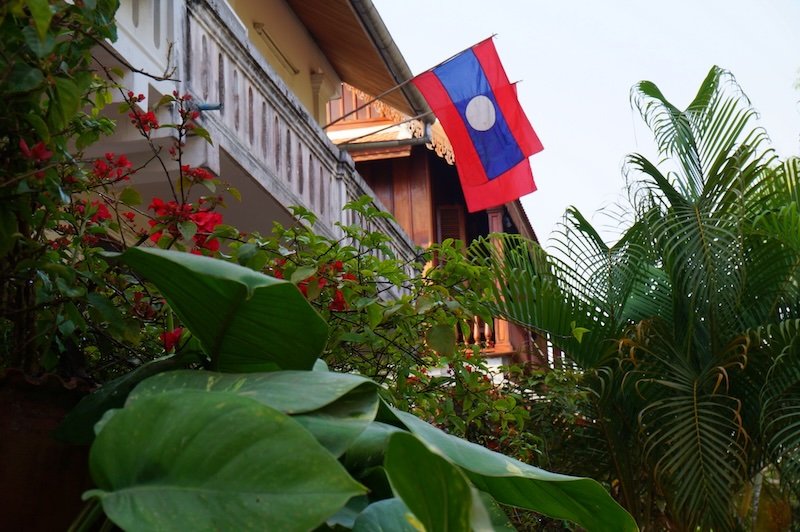 The national flag of Laos waves proudly from the balcony of a traditional wooden house in Luang Prabang. Lush tropical plants and vibrant flowers frame the scene, capturing the serene atmosphere and architectural charm of this UNESCO-listed town