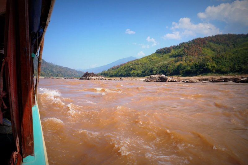 View from the side of a slow boat cruising down the muddy Mekong River in Laos, with swirling rapids in the foreground and lush green hills rising along the riverbanks under a clear blue sky during the scenic journey to Luang Prabang