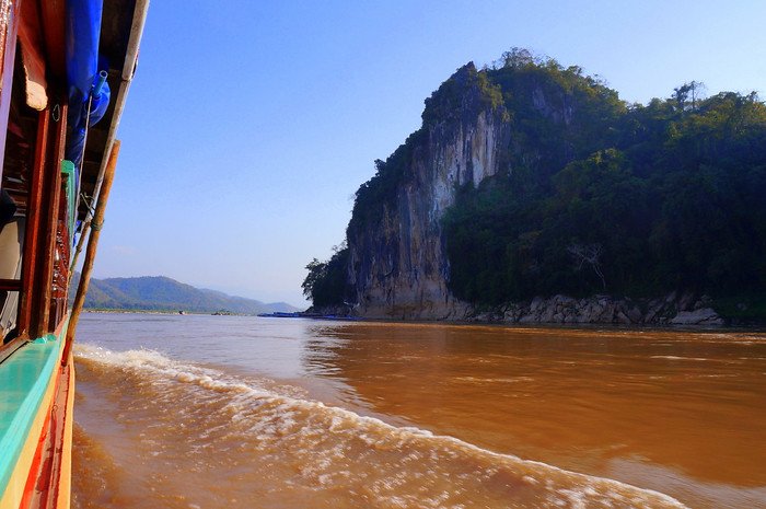 View from the slow boat on the Mekong River in Laos, passing towering limestone cliffs and riverbank caves on a sunny day. The muddy waters and dramatic rock formations create a striking landscape as the boat approaches Luang Prabang