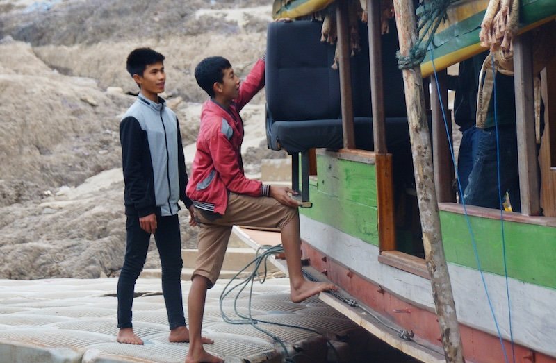 Two young men help load seats onto a traditional slow boat along the Mekong River in Laos. One balances barefoot on the dock while lifting the chair, capturing a candid glimpse of daily life and the practical preparations that go into river travel