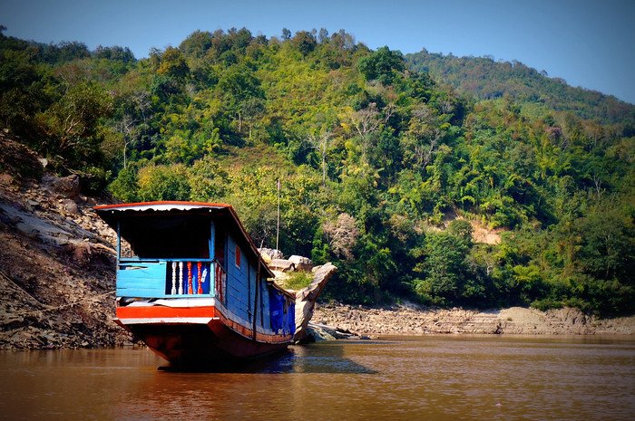 A traditional wooden slow boat painted blue and red moored along the Mekong River, surrounded by lush green hills in Laos. This scenic view captures the tranquil atmosphere experienced while traveling down the river en route to Luang Prabang