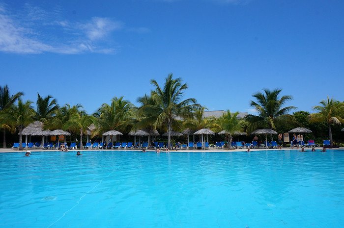Vacationers swim and lounge beneath swaying palm trees and palapa umbrellas around a large turquoise pool under clear blue skies at Melia Las Dunas resort in Cayo Santa María, Cuba, enjoying a sunny and relaxing honeymoon getaway