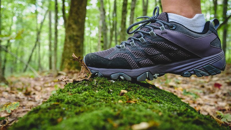 Close-up of a sturdy Merrell hiking shoe stepping on a moss-covered log along a forest trail in Sutton, Quebec, showcasing outdoor adventures in the Eastern Townships
