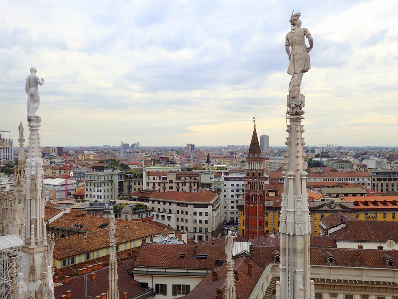 Panoramic cityscape of Milan, Italy viewed from the rooftop of Duomo di Milano, featuring intricate marble statues in the foreground and the red-brick bell tower of Chiesa di San Gottardo in Corte rising among the historic rooftops