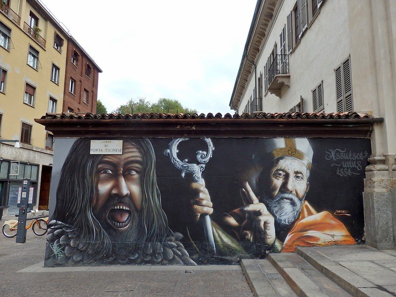 Vivid street art mural on Corso di Porta Ticinese in Milan, Italy, depicting a fierce warrior with flowing hair and a serene bishop holding a staff, showcasing the city’s vibrant urban art scene