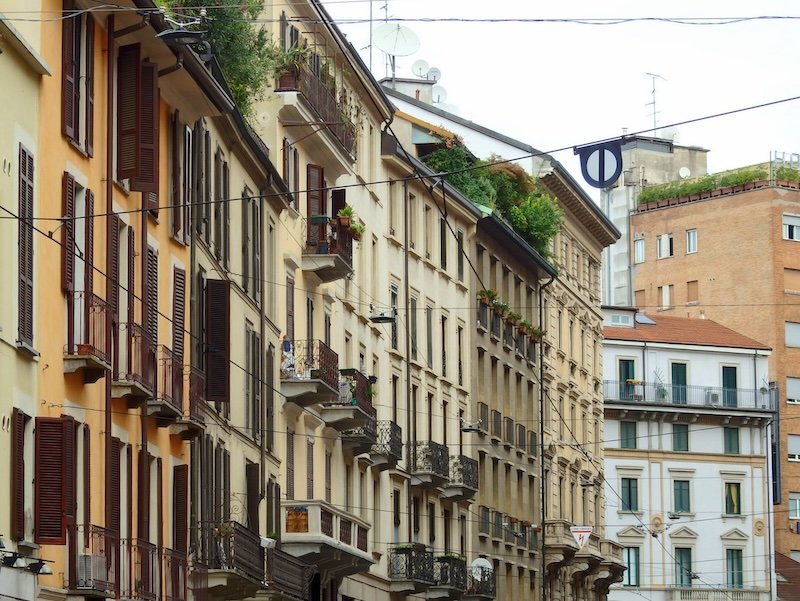 Row of classic residential buildings with wooden shutters, wrought-iron balconies, and rooftop greenery in Milan, Italy, showcasing the city’s elegant urban architecture