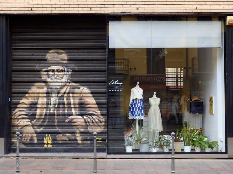 Street art mural of an elderly man in a hat painted on a storefront shutter beside a chic boutique window displaying elegant dresses and plants in Milan, Italy — blending urban art with high fashion in the city’s creative district