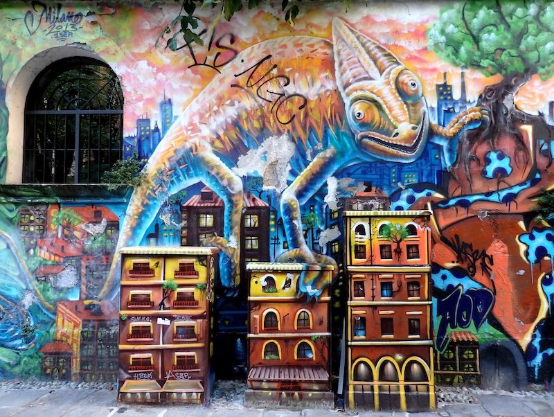 Colorful street art mural of a giant chameleon towering over painted city buildings on a vibrant urban wall in Milan, Italy, showcasing the city’s creative street scene