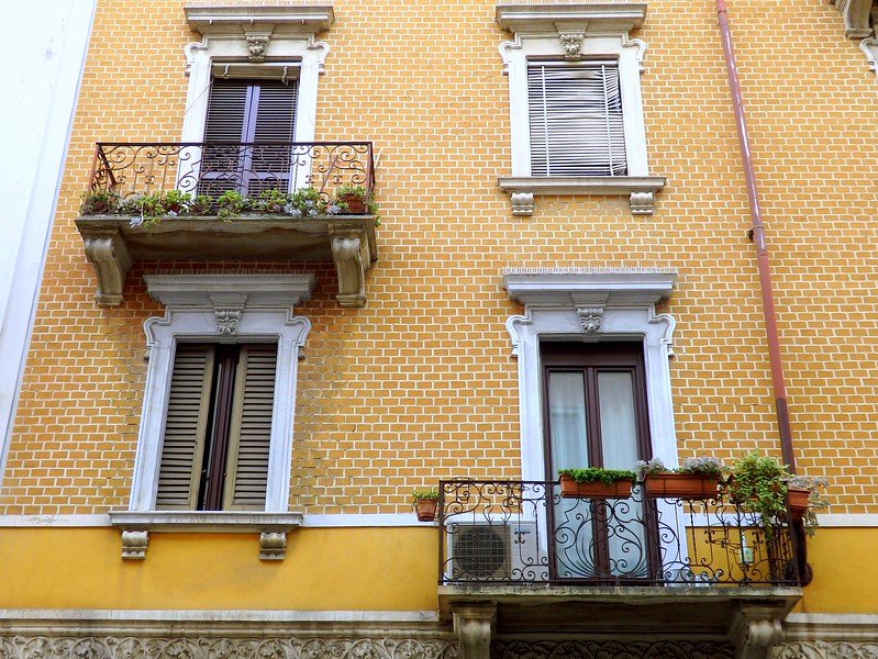 Charming yellow-brick residential building with ornate wrought-iron balconies and flower boxes in Milan, Italy, showcasing the city’s classic architectural style