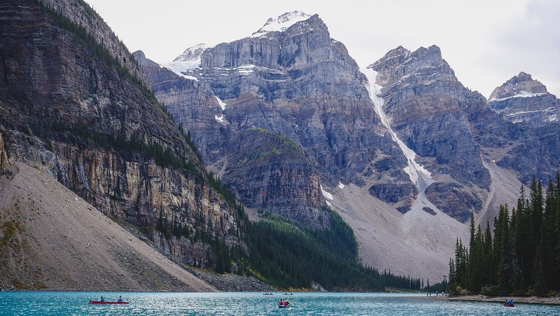 Towering snowcapped peaks and rugged cliffs rising above the turquoise waters of Moraine Lake, with small canoes paddling across the lake in the heart of the Canadian Rockies