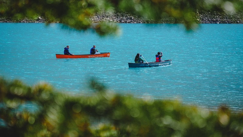 Two canoes gliding across the vivid turquoise-blue waters of Moraine Lake, framed by blurred evergreen branches, highlighting the lake’s striking electric colour in the Canadian Rockies