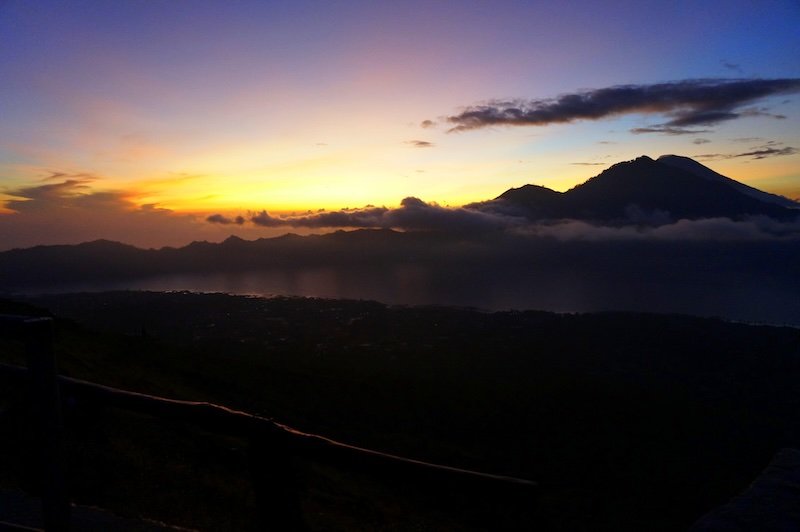 Golden sunrise breaking through the clouds above the silhouette of Mount Batur in Bali, Indonesia, casting warm light over the dark volcanic slopes and misty valleys below