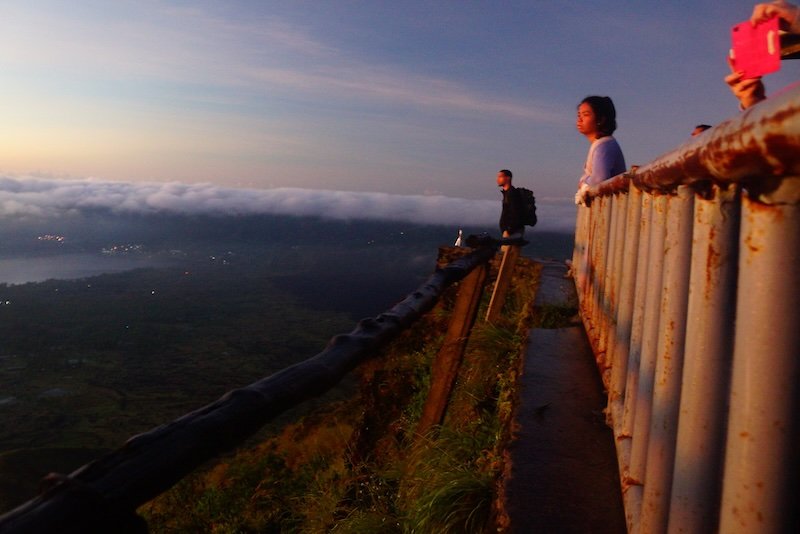 Travelers standing at the summit of Mount Batur in Bali, Indonesia, gazing over a sea of clouds and the green valley below at sunrise from a fenced lookout point