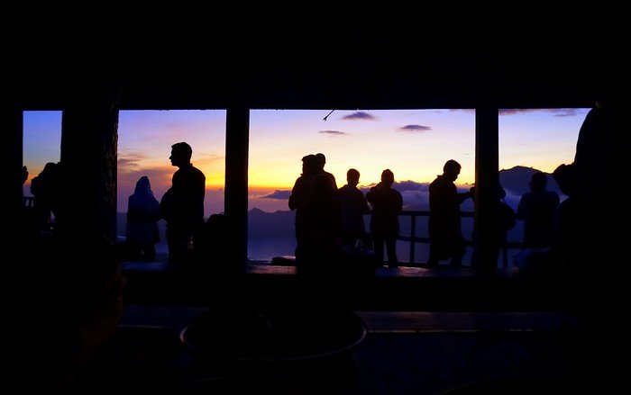 Silhouettes of hikers gathered at a summit hut on Mount Batur, watching the first light of dawn spread over the volcanic landscape of Bali, Indonesia