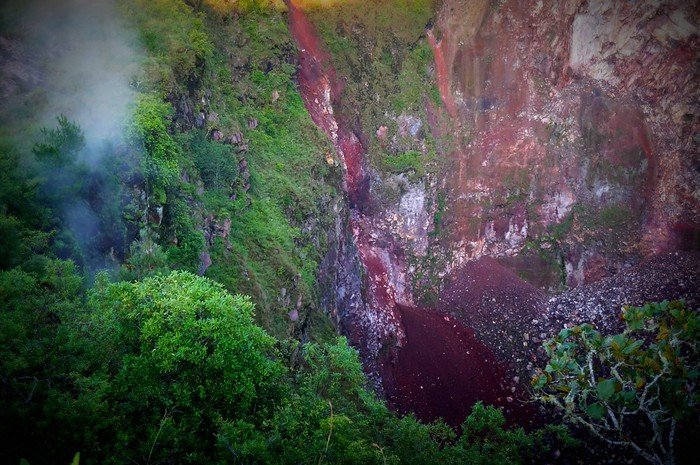 Steam rising from the rugged crater of Mount Batur in Bali, Indonesia, revealing layers of red volcanic rock and lush greenery clinging to the rim