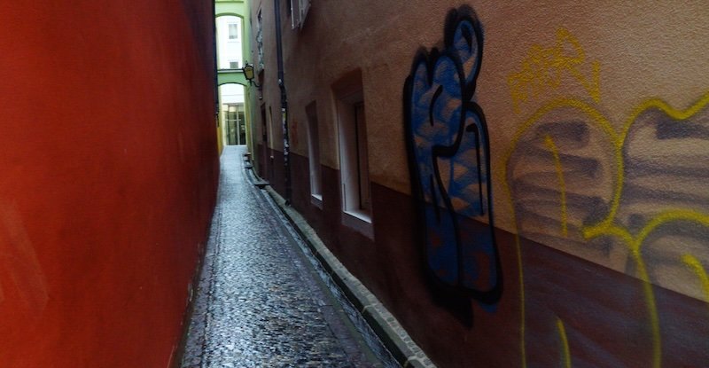 Narrow cobblestone alleyway in Freiburg im Breisgau framed by colourful walls with graffiti art, leading toward a bright passageway at the end