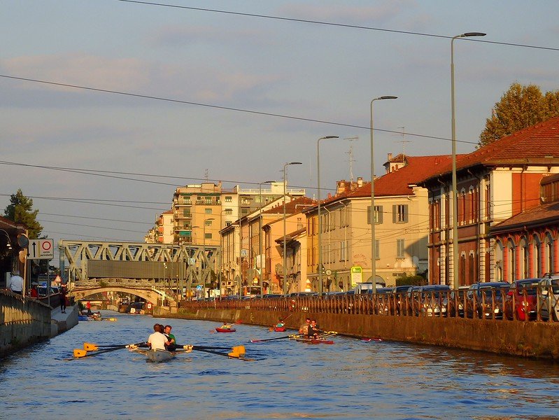 Rowers gliding along the Navigli canal at sunset in Milan, Italy, surrounded by historic pastel buildings, iron bridges, and lively waterfront streets bathed in golden light