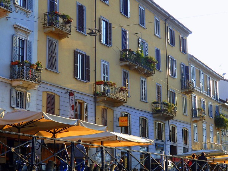 Charming pastel buildings with shuttered windows and flower-filled balconies overlooking lively canal-side cafes and outdoor dining terraces in the Navigli district of Milan, Italy, glowing in the golden afternoon light