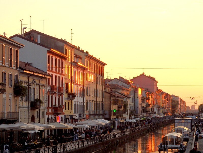 Golden hour in Milan, Italy, illuminating the colorful buildings and bustling cafés along the Navigli District canal, with reflections shimmering on the water as locals stroll the promenade