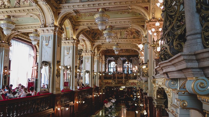 The lavish interior of the New York Café in Budapest, Hungary, featuring ornate gold detailing, grand chandeliers, arched ceilings, and elegant seating. Visitors enjoy coffee and pastries in this historic, must-visit landmark known as the “most beautiful café in the world.”