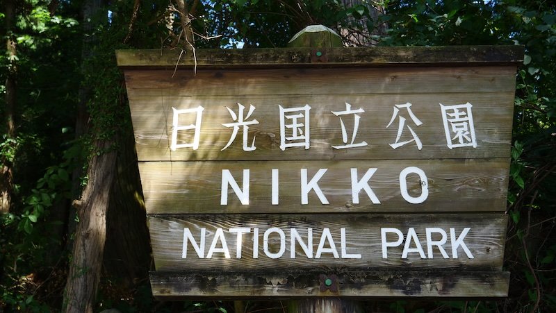 Weathered wooden entrance sign for Nikko National Park in Nikko, Japan, with white Japanese and English lettering set against a backdrop of dense forest greenery