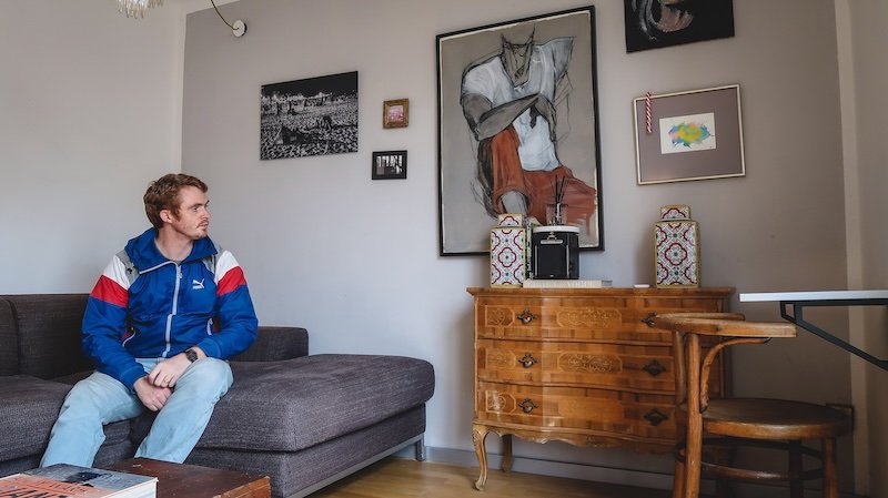 Nomadic Samuel sitting on a grey sofa inside a stylish Vienna Airbnb apartment, surrounded by eclectic wall art, a vintage wooden dresser, and cozy decor