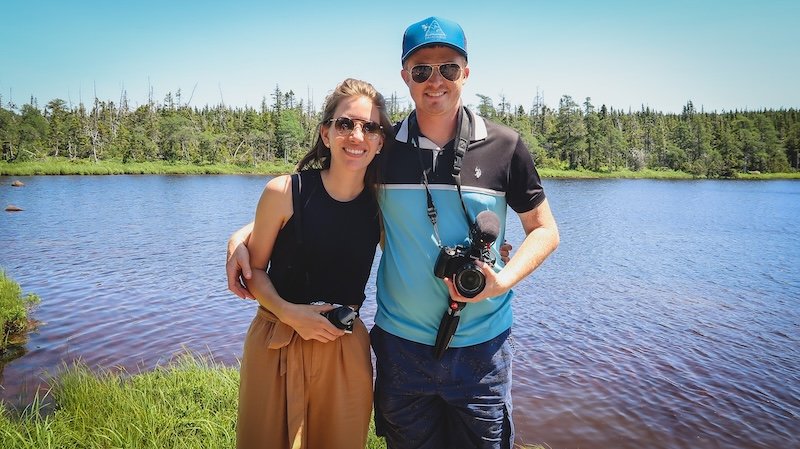 Nomadic Samuel and Audrey Bergner from That Backpacker smile happily while standing arm in arm by the calm waters of Benjie Lake in Cape Breton Highlands National Park, enjoying a sunny summer day on their scenic road trip along the Cabot Trail in Nova Scotia