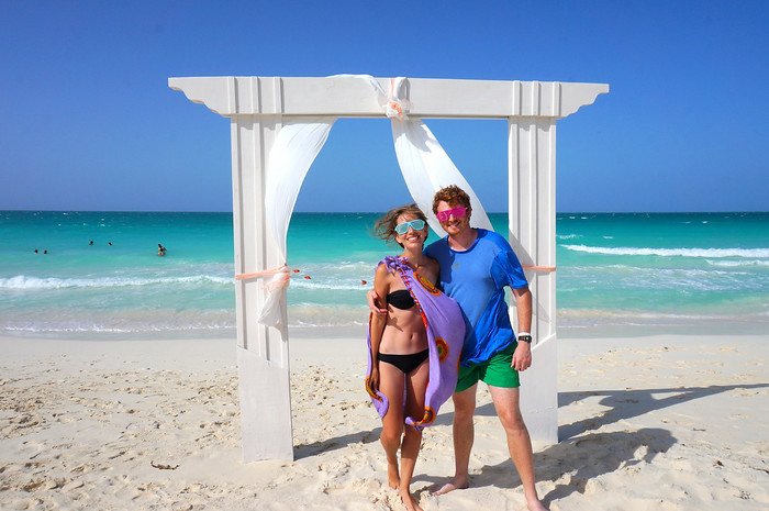 Nomadic Samuel and Audrey Bergner from That Backpacker smile as newlyweds under a white beach arch with flowing fabric, standing barefoot on the soft white sand with turquoise waves behind them during their honeymoon in Cayo Santa María, Cuba