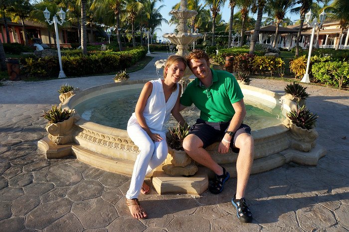Nomadic Samuel and Audrey Bergner from That Backpacker pose happily together by an elegant stone fountain framed by palm trees, tropical flowers, and pastel resort buildings during their romantic honeymoon at Cayo Santa María, Cuba