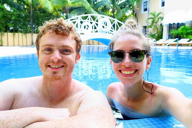 Nomadic Samuel and Audrey Bergner of That Backpacker smiling while relaxing in a resort pool during their one week in Playa del Carmen, Mexico, surrounded by tropical palm trees and sun loungers