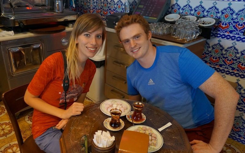 Nomadic Samuel and That Backpacker Audrey Bergner enjoying traditional Turkish tea served in tulip-shaped glasses with a plate of colorful Turkish delight at a cozy tiled café in Istanbul, Turkey, smiling and relaxed during their travels