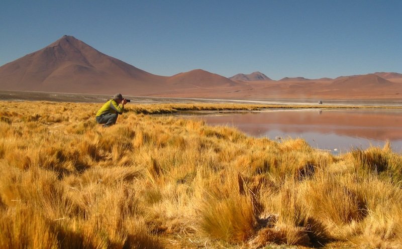 Nomadic Samuel crouching with camera during Bolivia salt flats 4x4 tour, capturing Andean landscapes and flamingo lagoon