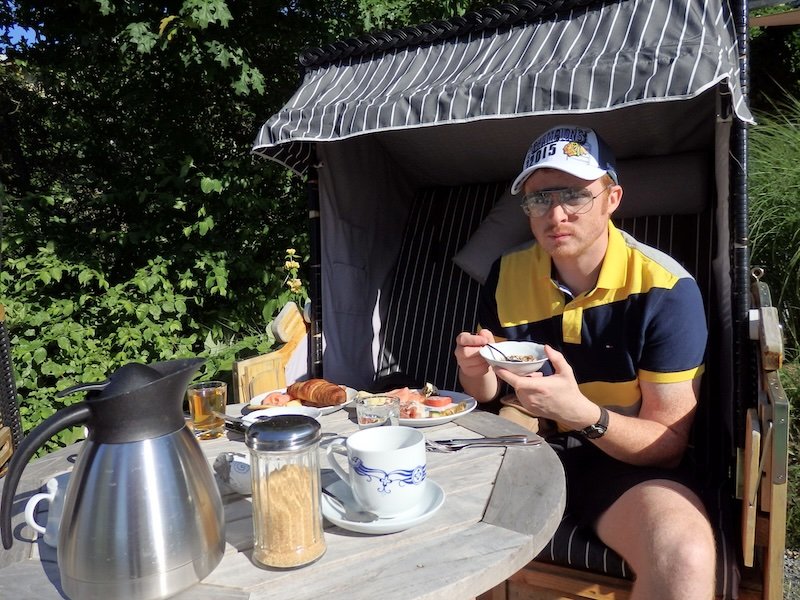 Nomadic Samuel enjoying a leisurely breakfast outdoors on Rügen Island, Germany, seated in a traditional Strandkorb beach chair with a table set with coffee, croissants, cold cuts, and fresh fruit under the morning sun