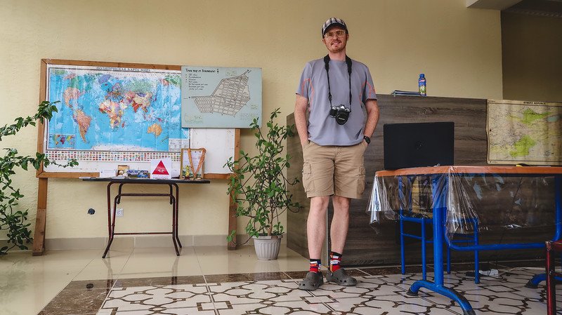 Nomadic Samuel standing with a camera around his neck at the CBT Bokonbayevo office in Kyrgyzstan, surrounded by world and regional maps before beginning the 3-Day Panorama Trek