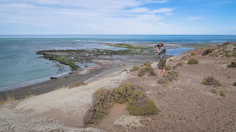 Nomadic Samuel stands on a sandy cliff overlooking the rugged coastline of Puerto Pirámides, Península Valdés, Argentina, capturing the dramatic seascape with his camera. The scene highlights tidal flats, rocky outcrops, and sweeping ocean views