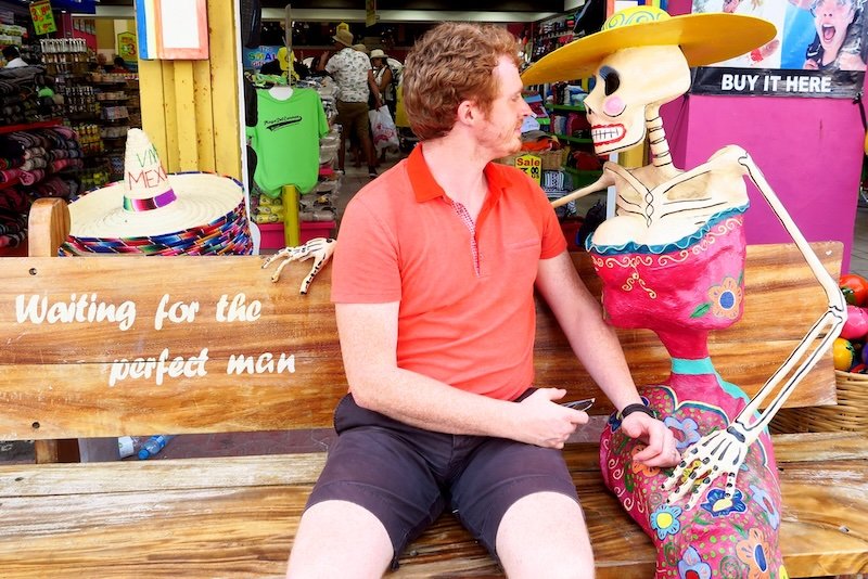Nomadic Samuel sitting on a souvenir shop bench in Playa del Carmen beside a colorful skeleton figure wearing a sombrero, creating a humorous travel moment Nomadic Samuel sitting on a souvenir shop bench in Playa del Carmen beside a colorful skeleton figure wearing a sombrero, creating a humorous travel moment