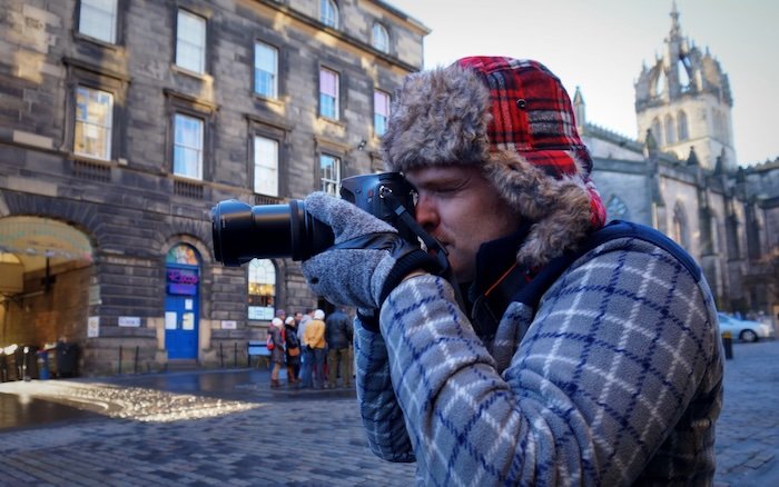 Nomadic Samuel dressed for winter weather in Edinburgh, Scotland wearing a warm hat and taking photos at street level 