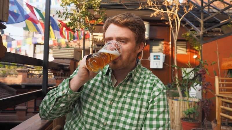 Nomadic Samuel enjoying a cold beer on a rooftop terrace in Kathmandu, Nepal, with prayer flags and local city views in the background