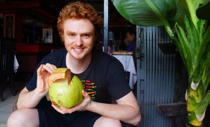 Nomadic Samuel smiling while holding a freshly opened green coconut at a café in Siem Reap, Cambodia during his one-month Cambodia travel itinerary