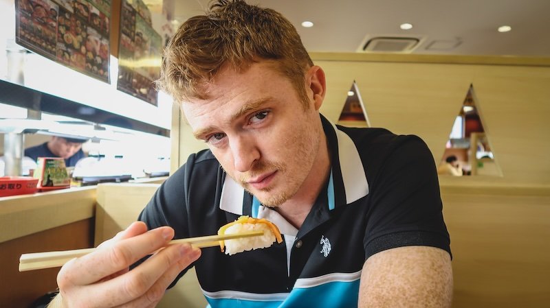 Nomadic Samuel eating conveyor belt sushi in Osaka, Japan with chopsticks 
